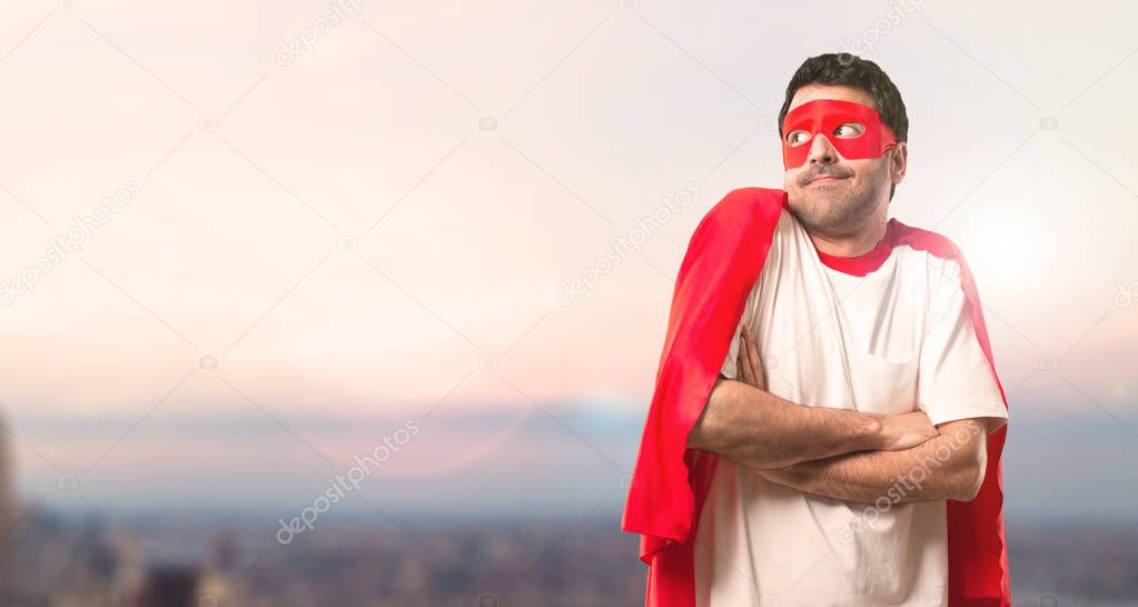 Superhero man with mask and red cape making unimportant and doubts gesture while lifting the shoulders on a sunset background