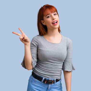 Gülümseyen ve zafer işareti mavi arka plan üzerinde gösterilen genç Kızıl saçlı kız