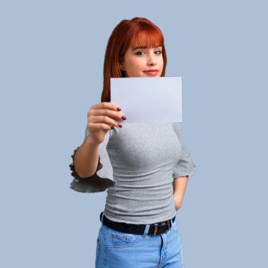 Boş bir afişini mavi arka plan üzerinde tutan genç Kızıl saçlı kız