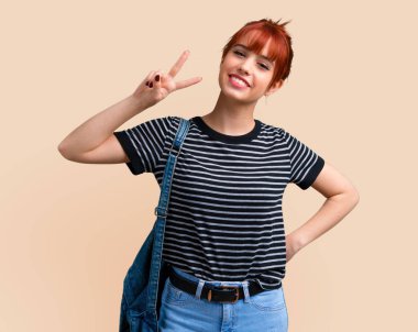 Gülümseyen ve zafer işareti koyu sarı arka plan üzerinde gösterilen genç öğrenci Kızıl saçlı kız