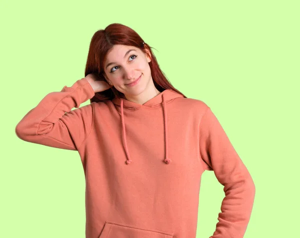 粉红色运动衫的年轻红发女孩站在和思考一个想法 而抓头在绿色背景 — 图库照片