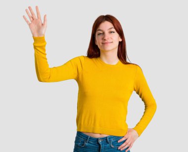 Gri arka plan üzerinde mutlu ifade ile el ile sarı kazak selamlayan ile genç Kızıl saçlı kız