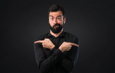 Handsome man with beard pointing to the laterals having doubts on black background clipart