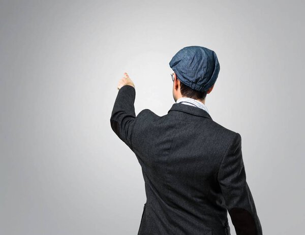 Handsome modern man with beret and glasses pointing back with the index finger on grey background