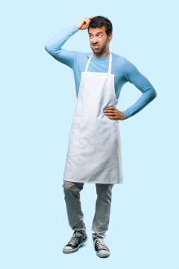 Tam vücut şüpheleri bir önlük giyen adam ve tırmalamak baş üzerinde mavi arka planı süre karıştırmayın yüz ifadesi ile