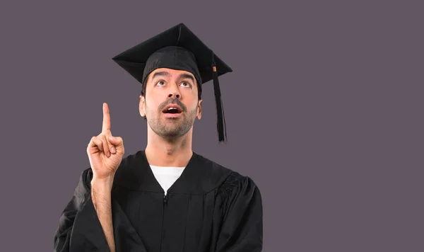 立っていると 紫色の背景に指を上向きのアイデアを考えて彼の卒業日大学の男 — ストック写真