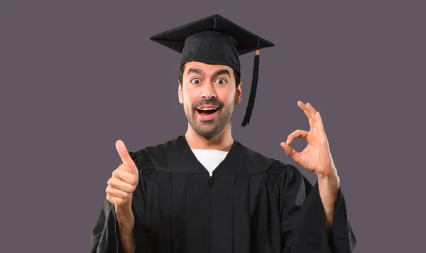 彼の卒業の日の大学指で サインを示し もう一方の手でジェスチャーを親指を与える紫色の背景の人 — ストック写真