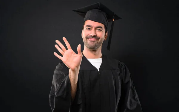 Man on his graduation day University saluting with hand with happy expression on black background