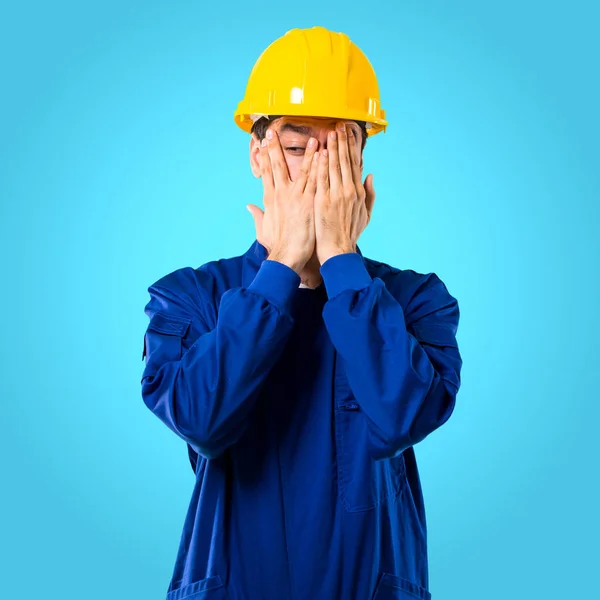 年轻的工人与头盔覆盖眼睛的手和看通过手指在蓝色背景 — 图库照片