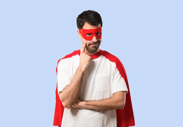 Superhero man with mask and red cape standing and looking to the front opening the eye with finger on isolated blue background
