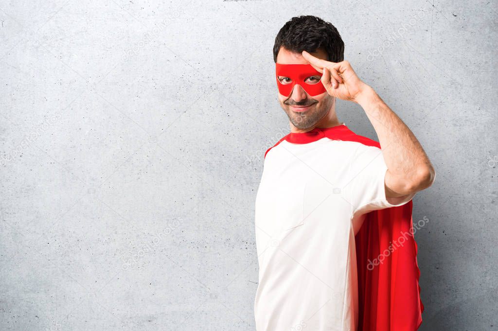 Superhero man with mask and red cape saluting with hand with happy expression on textured grey background