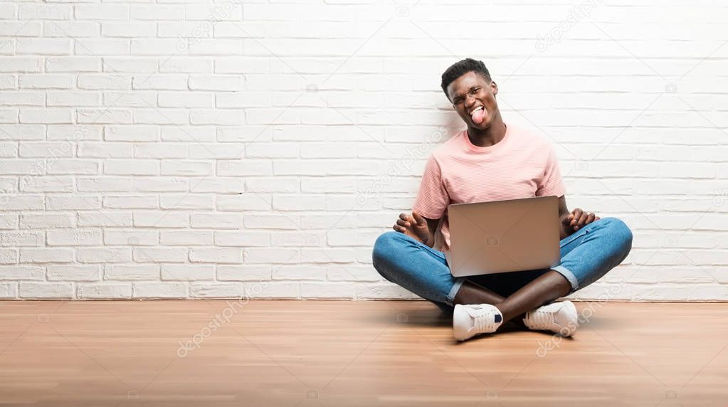 Afro american man sitting on the floor with his laptop showing tongue at the camera having funny look