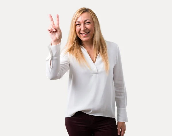 笑みを浮かべて 孤立した灰色の背景に陽気な顔に勝利のサインを示す白いシャツ中年金髪女性 — ストック写真