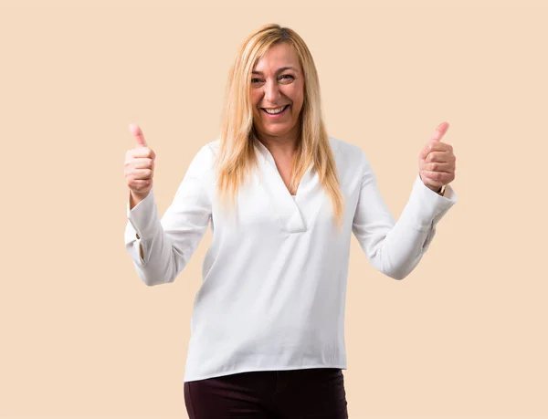 Middle age blonde woman with white shirt giving a thumbs up gesture with both hands and smiling. Cheerful expression on isolated ocher background