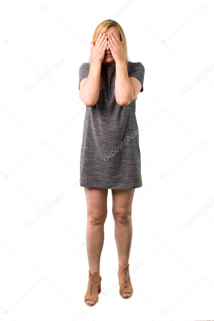 Middle age blonde woman with grey dress covering eyes by hands. Do not want to see something on isolated white background