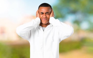 Her iki kulak elleriyle kapsayan beyaz kazak olan genç adam koyu tenli. Bir parkta hayal kırıklığı ifade