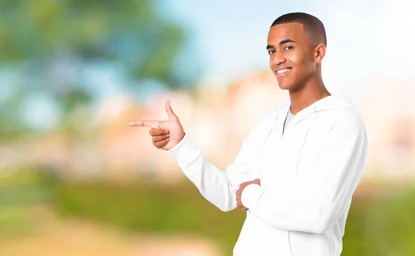深色皮肤的年轻男子 白色运动衫指着手指侧面 并提出了一个产品在一个公园的横向位置 — 图库照片