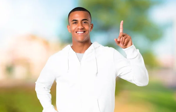 黑皮肤的年轻人与白色运动衫显示和举起手指在公园最好的标志 — 图库照片