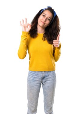 Sarı kazak ve parmaklar ok işaretiyle gösterilen ve izole beyaz arka plan üzerinde jest diğer elinizle bir başparmak veren kafasındaki mavi bandana ile genç kız