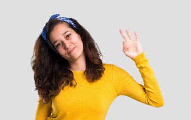 Sarı kazak ve parmaklar ok işaretiyle gösterilen başında mavi bandana ile genç kız. Mutluluk ve memnuniyet izole gri arka plan üzerinde yüzü