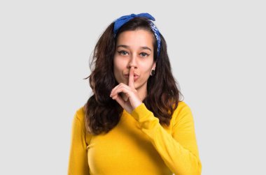 Sarı kazak ve kafasındaki bir işaret parmağını ağzına izole gri arka plan üzerine koyarak ağız ve sessizlik bir jest kapanış gösteren mavi bandana ile genç kız