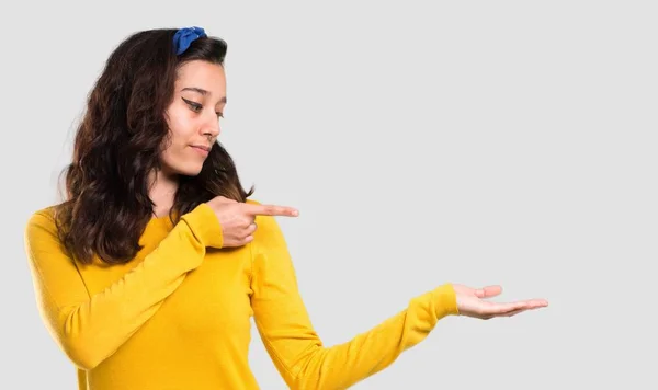 年轻的女孩与黄色毛衣和蓝色头巾在她的头持有 Copyspace 假想在手掌上插入广告在孤立的灰色背景 — 图库照片