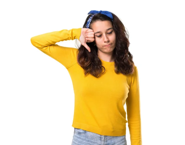 黄色いセーターを着て 彼女の頭を親指で下を負の式と記号を示す青いバンダナを持つ少女 孤立した白い背景の悲しそうな表情 — ストック写真