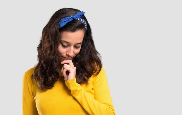 年轻的女孩与黄色毛衣和蓝色头巾在她的头站立和向下看与手在查出的灰色背景的下巴上 — 图库照片
