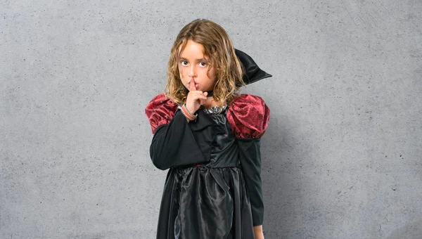 Kid dressed as a vampire at halloween holidays showing a sign of closing mouth and silence gesture on textured background