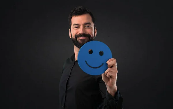 Handsome man with beard holding a happy face icon on black background