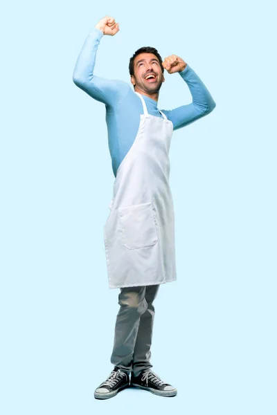 Full body of Man wearing an apron celebrating a victory and surprised to be successful on blue background