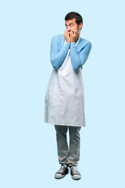 Full body of Man wearing an apron is a little bit nervous and scared putting hands to mouth on blue background