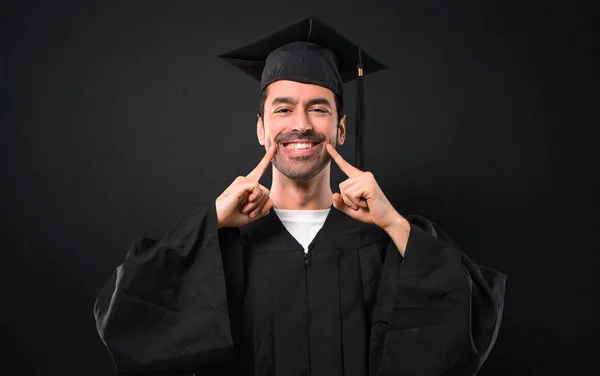 笑みを浮かべて彼卒業日大学黒い背景に口と指で顔を指差しながら幸せと楽しい式を持つ男 — ストック写真