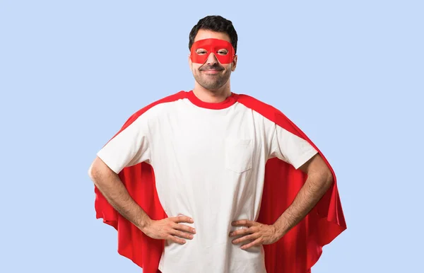 Superhero man with mask and red cape posing with arms at hip and laughing looking to the front on isolated blue background