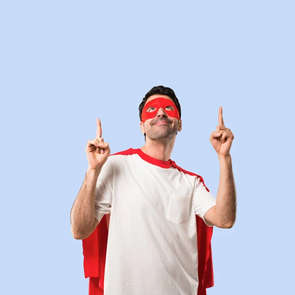 Superhero man with mask and red cape pointing with the index finger a great idea and looking up on isolated blue background