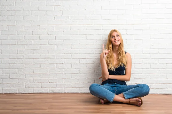 白いレンガ壁の背景に素晴らしいアイデア人差し指で指している床の上に座ってブロンドの女の子 — ストック写真