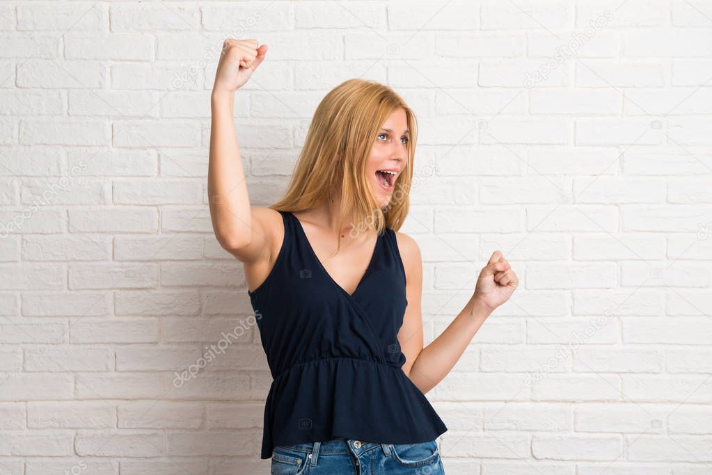 Blonde woman celebrating a victory and happy for having won a prize on white brick wall background