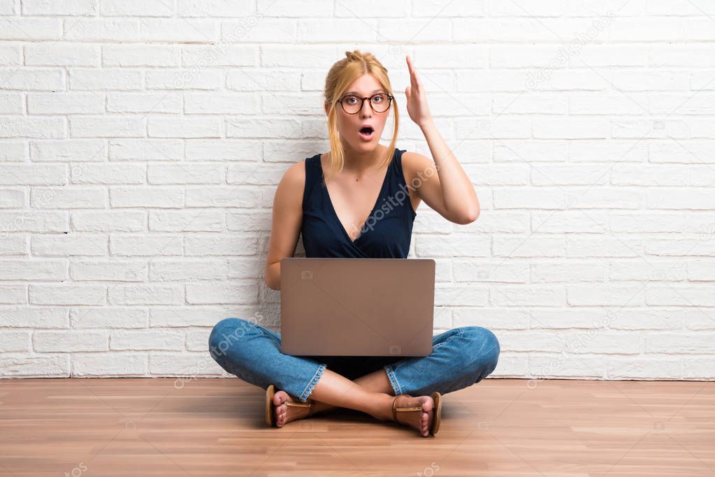 Blonde girl sitting on the floor with her laptop intending to realizes the solution on white brick wall background