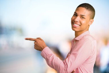 Parmak tarafına işaret ve odaklanmamış açık arka planda lateral pozisyonda bir ürün sunma genç Afro-Amerikan adam