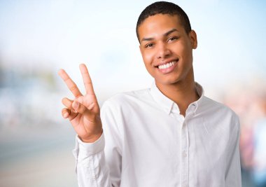 Gülümseyen ve neşeli bir yüz zafer işaretiyle odaklanmamış açık arka plan üzerinde gösterilen genç Afro-Amerikan adam