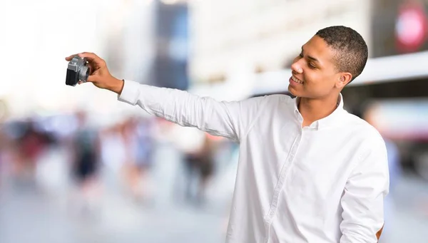 Young african american man with white shirt making selfie with old camera in the middle of the city