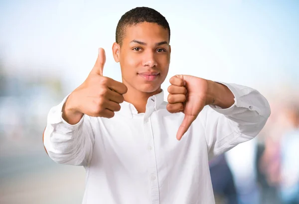 Young african american man with white shirt making good-bad sign. Undecided person between yes or not on unfocused outdoor background