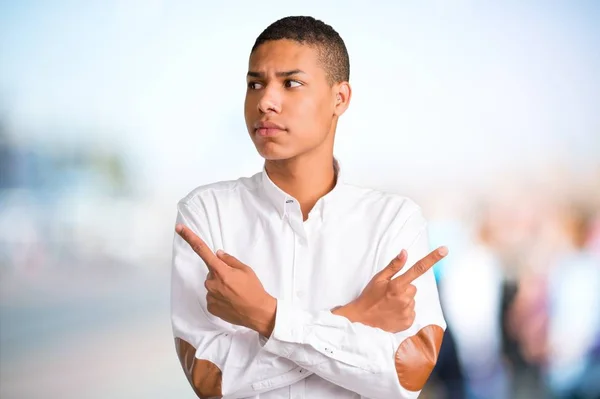年轻的非洲裔美国男子与白色衬衫指向的分支有疑虑 未决定的人在不聚焦的户外背景 — 图库照片