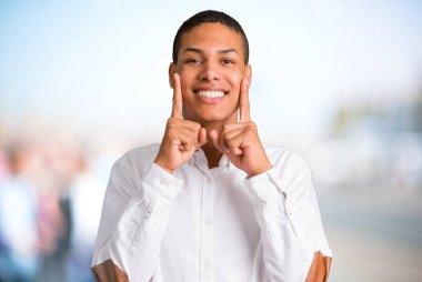 Beyaz gömlek odaklanmamış açık arka plan ağız ve yüz parmak ile işaret ederken mutlu ve keyifli bir ifade ile gülümseyen genç Afro-Amerikan adamla