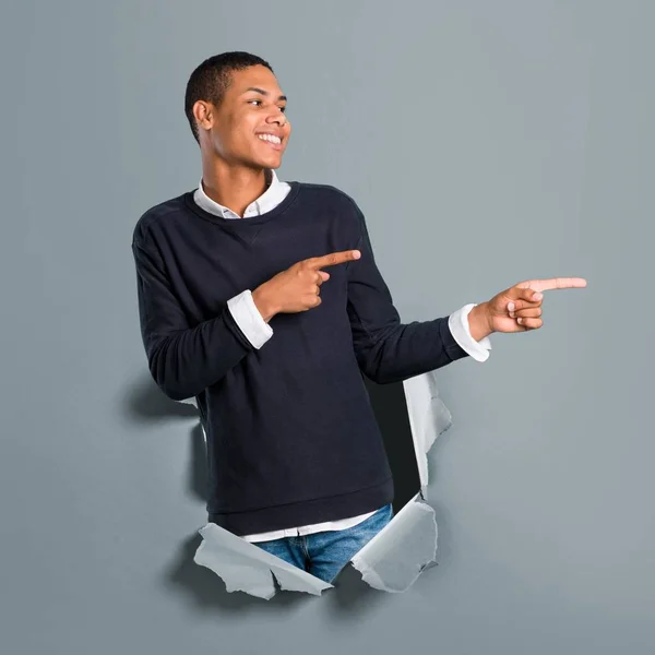 Young african american man pointing finger to the side and presenting a product while smiling through a paper hole