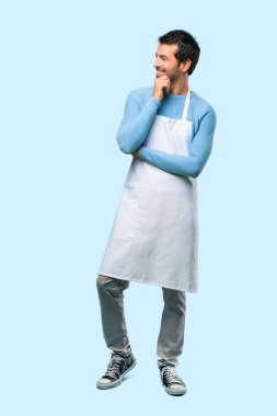 Ayakta ve el ile mavi arka plan çeneme tarafına bakıp bir önlük giyen adamın tam vücut