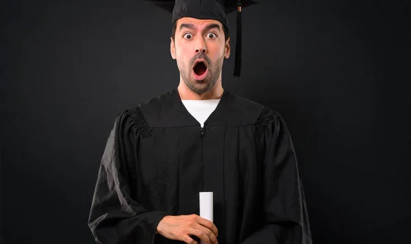 Man on his graduation day University with surprise and shocked facial expression. Gaping because have just surprised with a gift on black background