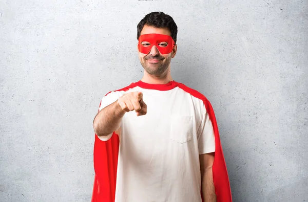 Superhero man with mask and red cape points finger at you with a confident expression on textured grey background