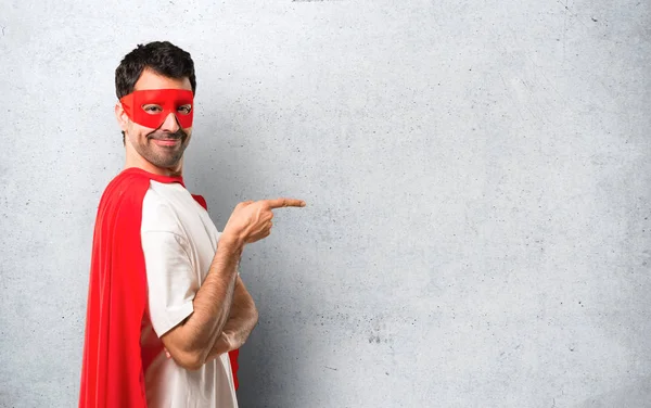 Superhero man with mask and red cape pointing finger to the side and presenting a product in lateral position on textured grey background