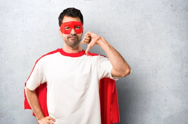 Superhero man with mask and red cape showing thumb down sign with negative expression. Sad expression on textured grey background